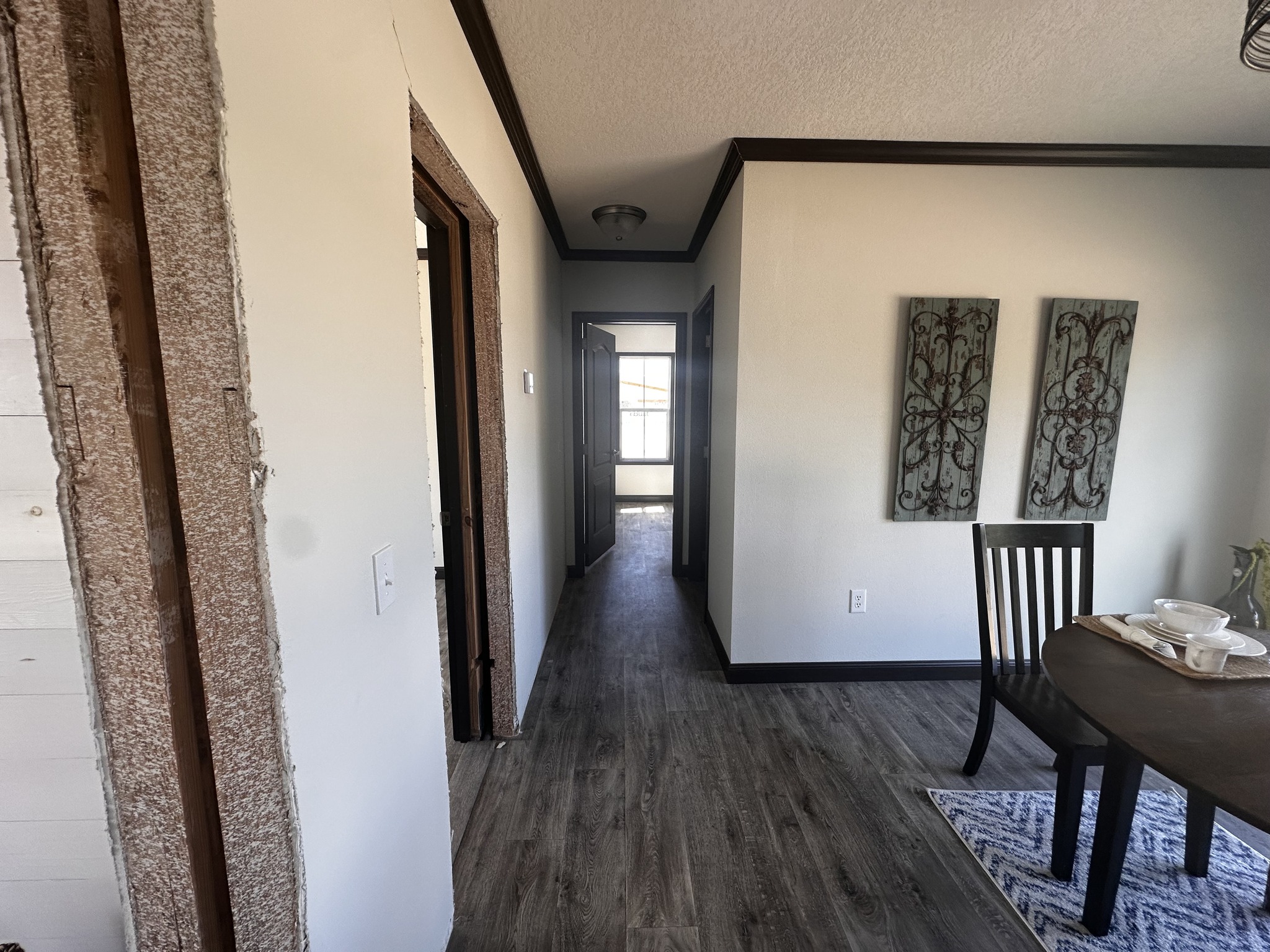 A hallway with dark wood flooring leads to a bright window. Decor on the left wall; a dining table with place settings on the right adds warmth.