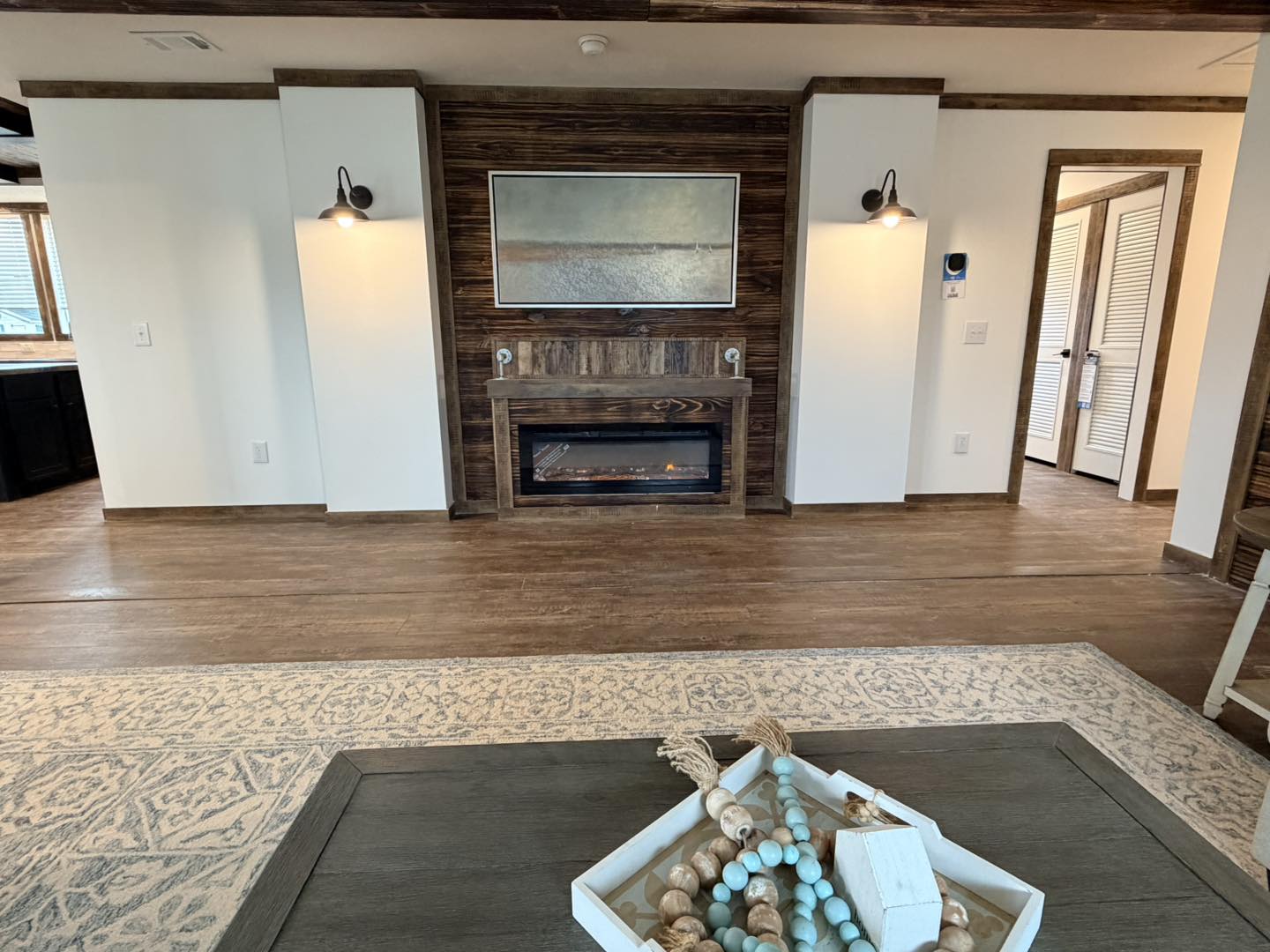 Modern living room with a wood-paneled electric fireplace, a painting above it, and two wall sconces. A decorative tray on a coffee table in the foreground.
