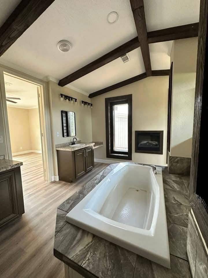 Luxurious bathroom with a white bathtub set in stone tiles, dark wood ceiling beams, a vanity with a mirror, and a window brightening the space.