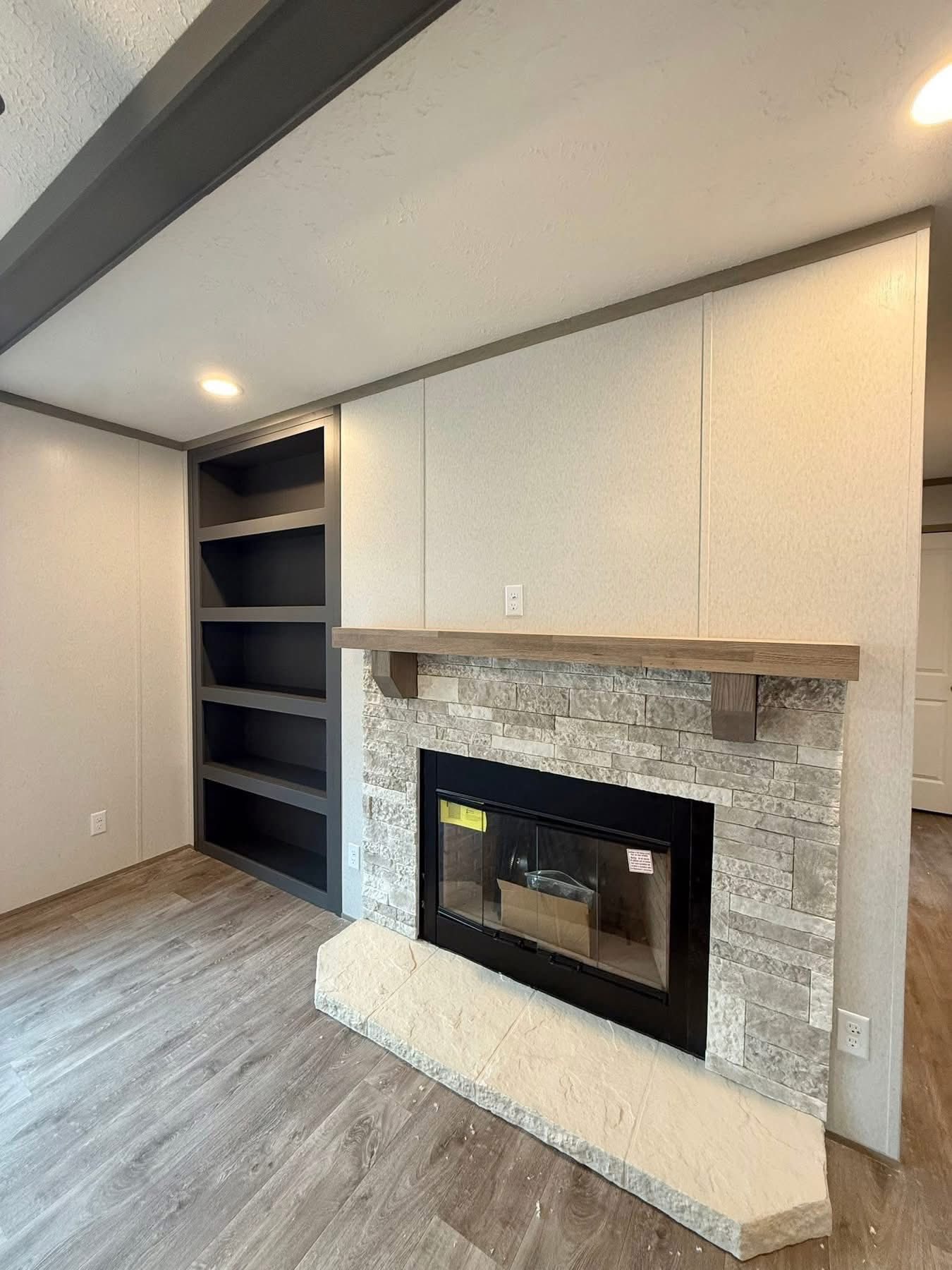 Modern living room with a stone fireplace and wooden mantel. Adjacent are dark, built-in shelves. The flooring is wood, creating a warm, cozy atmosphere.