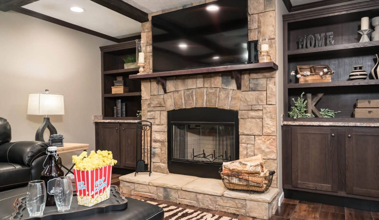 Cozy living room with a stone fireplace, dark wooden shelves, and a large TV above. A table with popcorn, drinks, and glasses adds a relaxed, inviting feel.