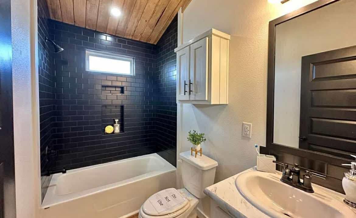 Cozy bathroom with black tile bathtub, wooden ceiling, and small window. Modern white sink, cabinet, toilet, and potted plant create a warm, inviting tone.
