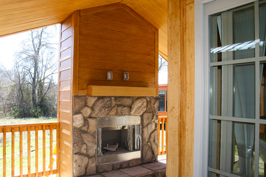 Covered porch with a rustic stone fireplace. Wooden paneling and railing add warmth. Large windows and trees in the background enhance a cozy feel.