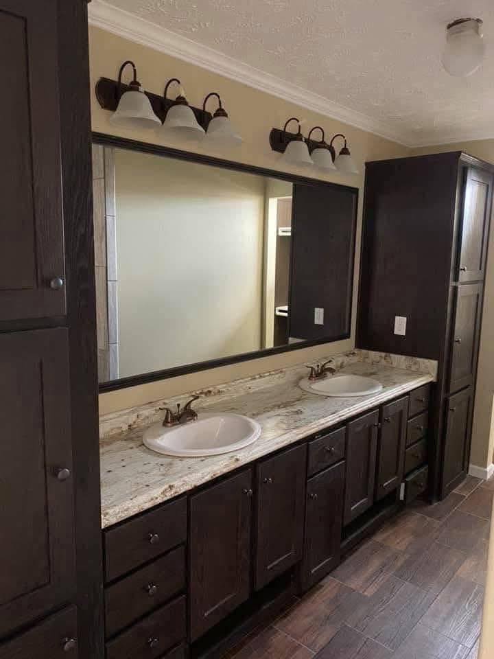 A modern bathroom features a double vanity with dark wood cabinets and a speckled marble countertop. Two white sinks, a large mirror, and warm lighting create an elegant and cozy atmosphere.