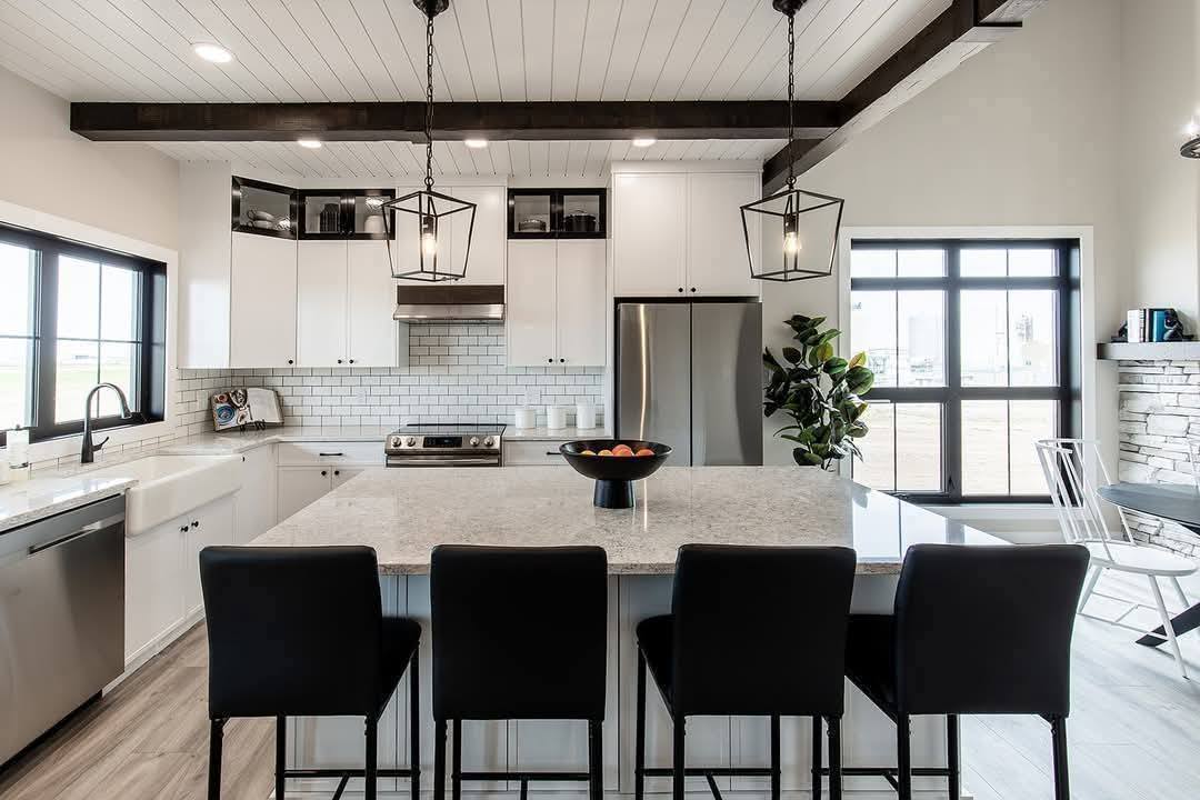 Modern kitchen with a large marble island, black chairs, stainless steel appliances, and white cabinetry. Bright lighting and a cozy atmosphere.