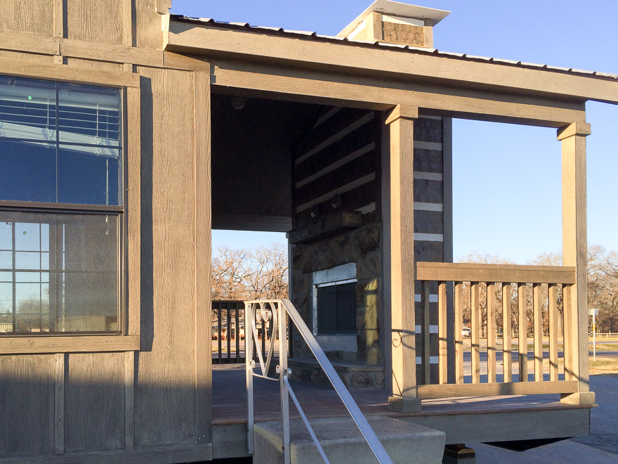 Wooden porch with railing attached to a rustic building. The porch features a stone fireplace and overlooks a sunlit, tree-filled landscape.