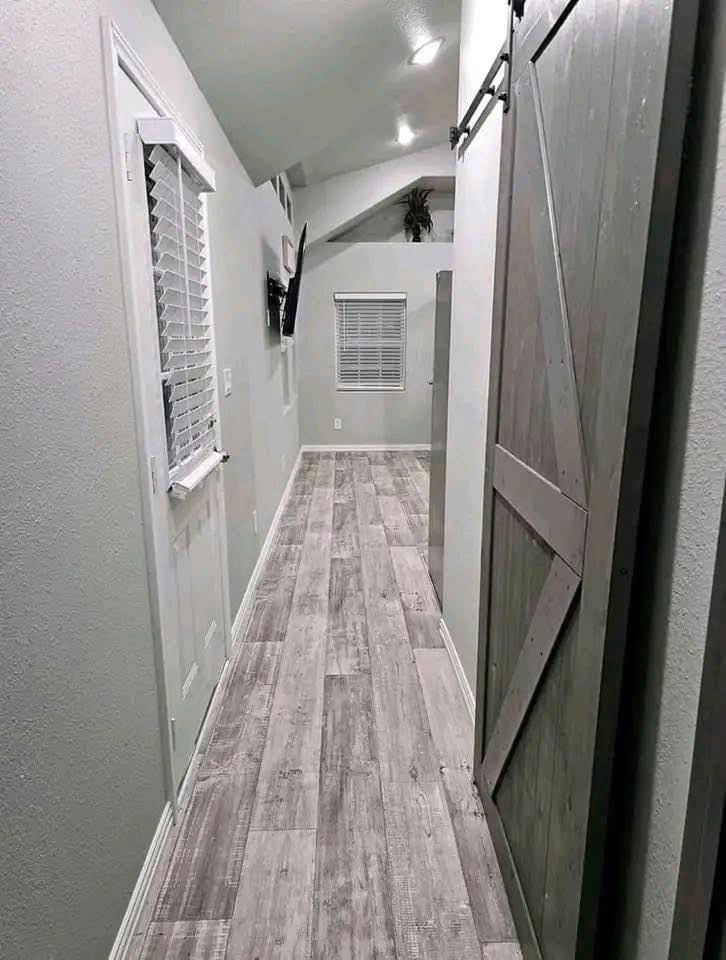Narrow hallway with grey wood flooring, flanked by white walls. A closed barn-style sliding door is on the right, window with blinds on the left.