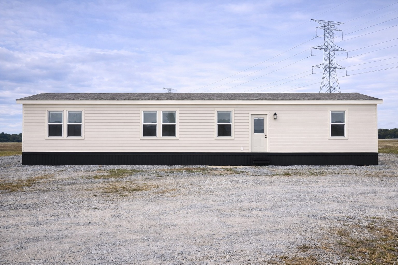 A beige modular home with five windows and a central door sits on a gravel lot. A tall electrical tower stands in the background under a cloudy sky.