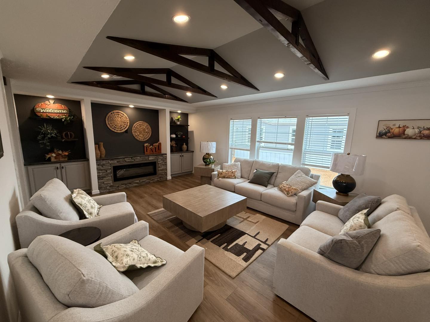 Stylish living room with light gray sofas around a wooden coffee table. Features exposed beams, large windows, and a modern fireplace, creating a cozy ambiance.