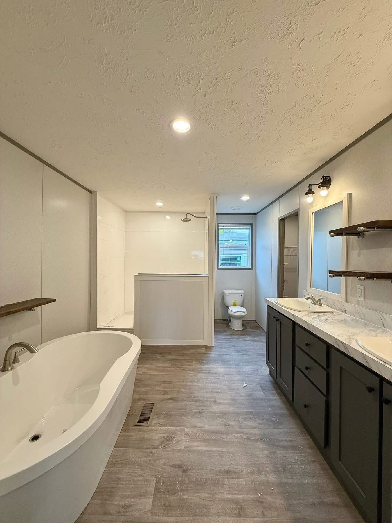 Spacious modern bathroom with a freestanding tub, double sinks on a marble countertop, dark cabinetry, a walk-in shower, and wood-patterned flooring.