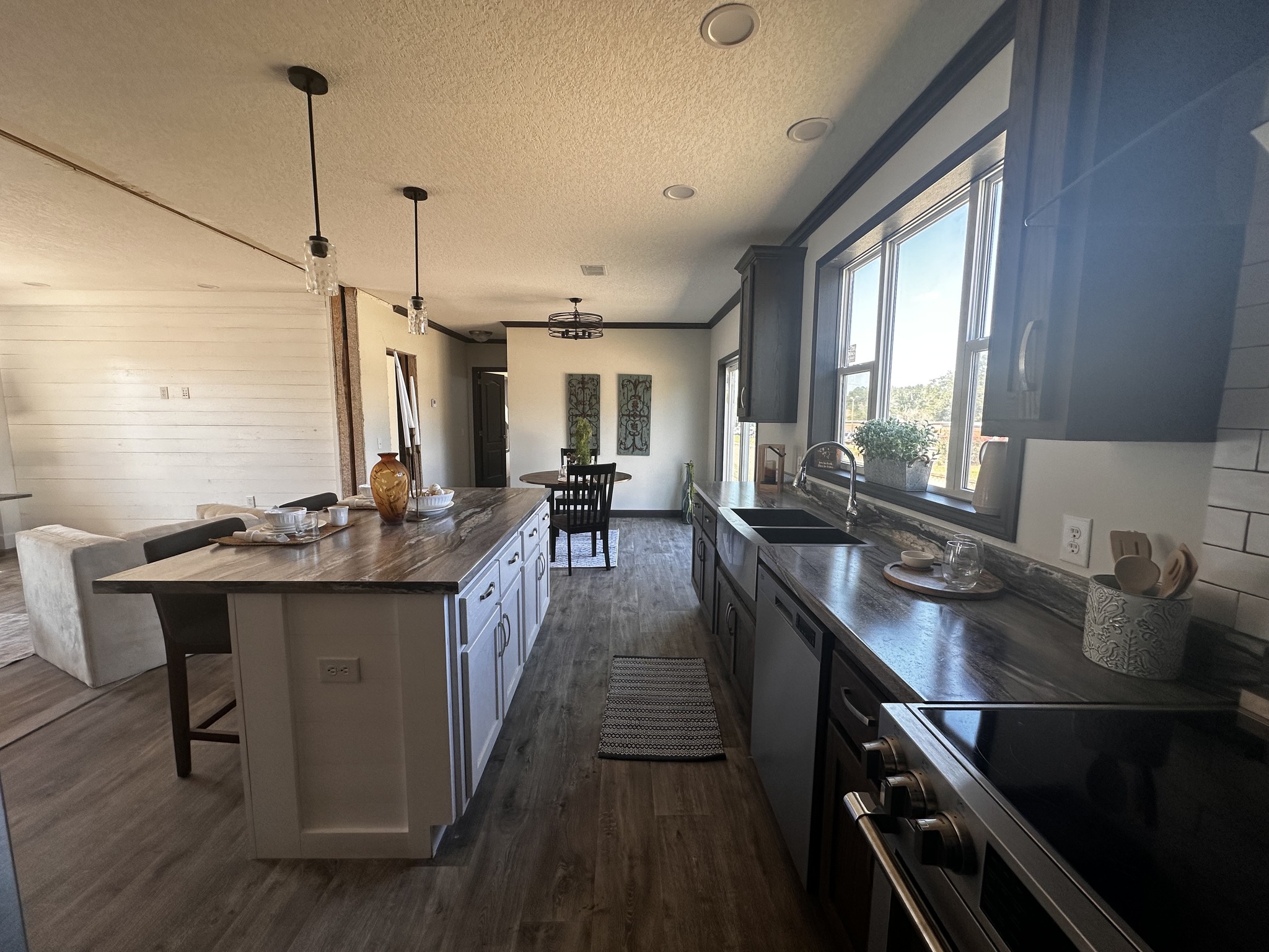 Modern kitchen with wood floors features a large island with bar stools, pendant lights, and a window-lit sink area. Dining table in background.