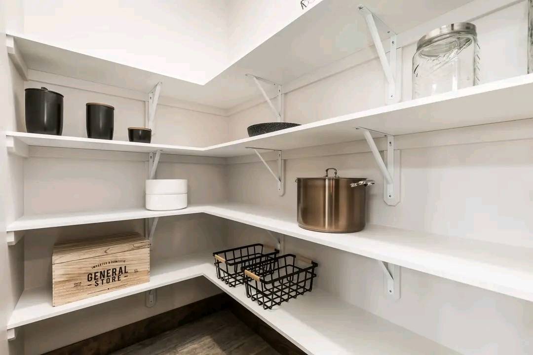 A neatly organized pantry with white shelves holding glass jars, a metal pot, black canisters, and woven baskets. A wooden box reads "General Store."