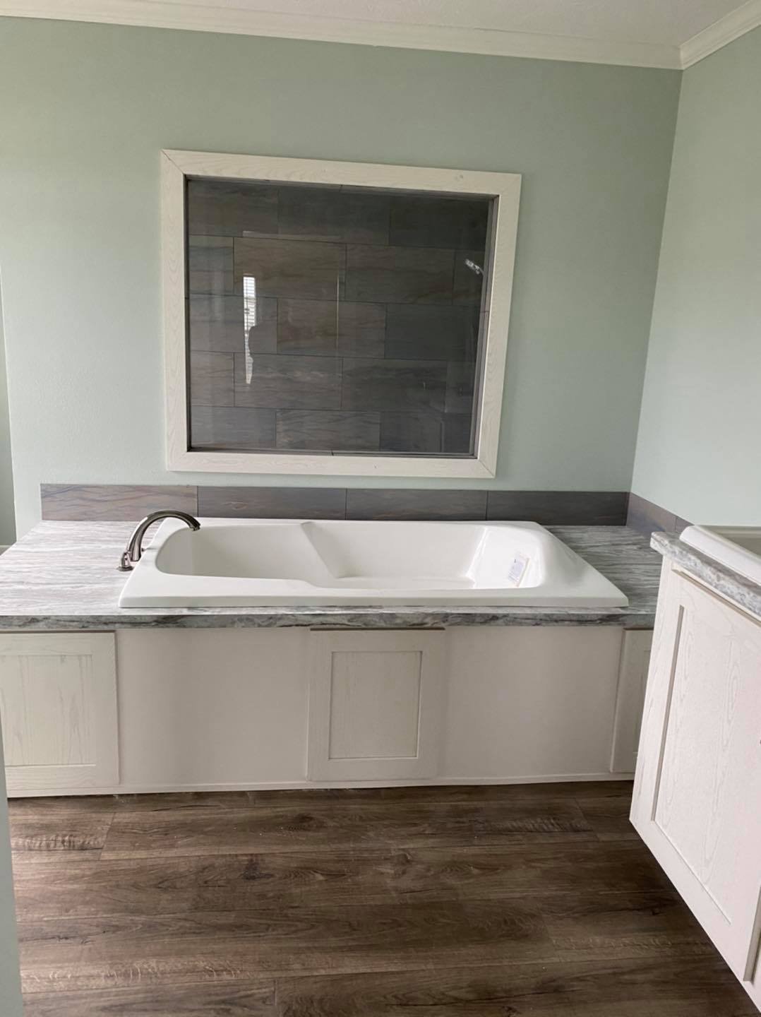 A white bathtub with a silver faucet is set in a gray marble surround. A large window overlooks the tub. The room features light green walls and wooden flooring, creating a calm ambiance.
