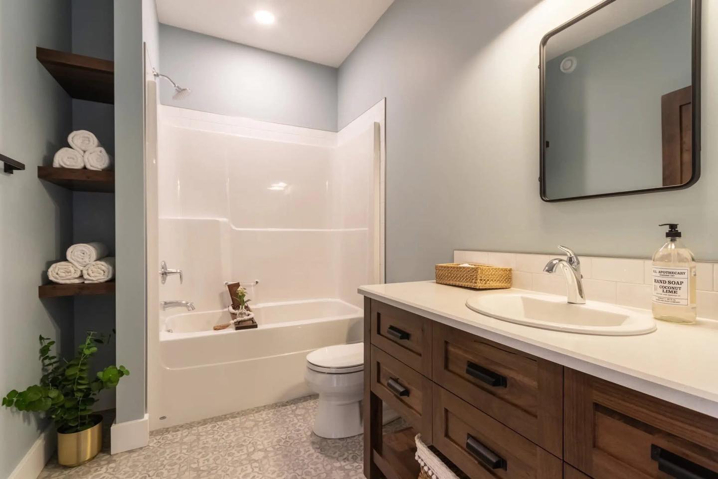 A modern bathroom with a sleek tub, wooden vanity, and square mirror. Shelves hold rolled towels and a potted plant, creating a clean, serene atmosphere.