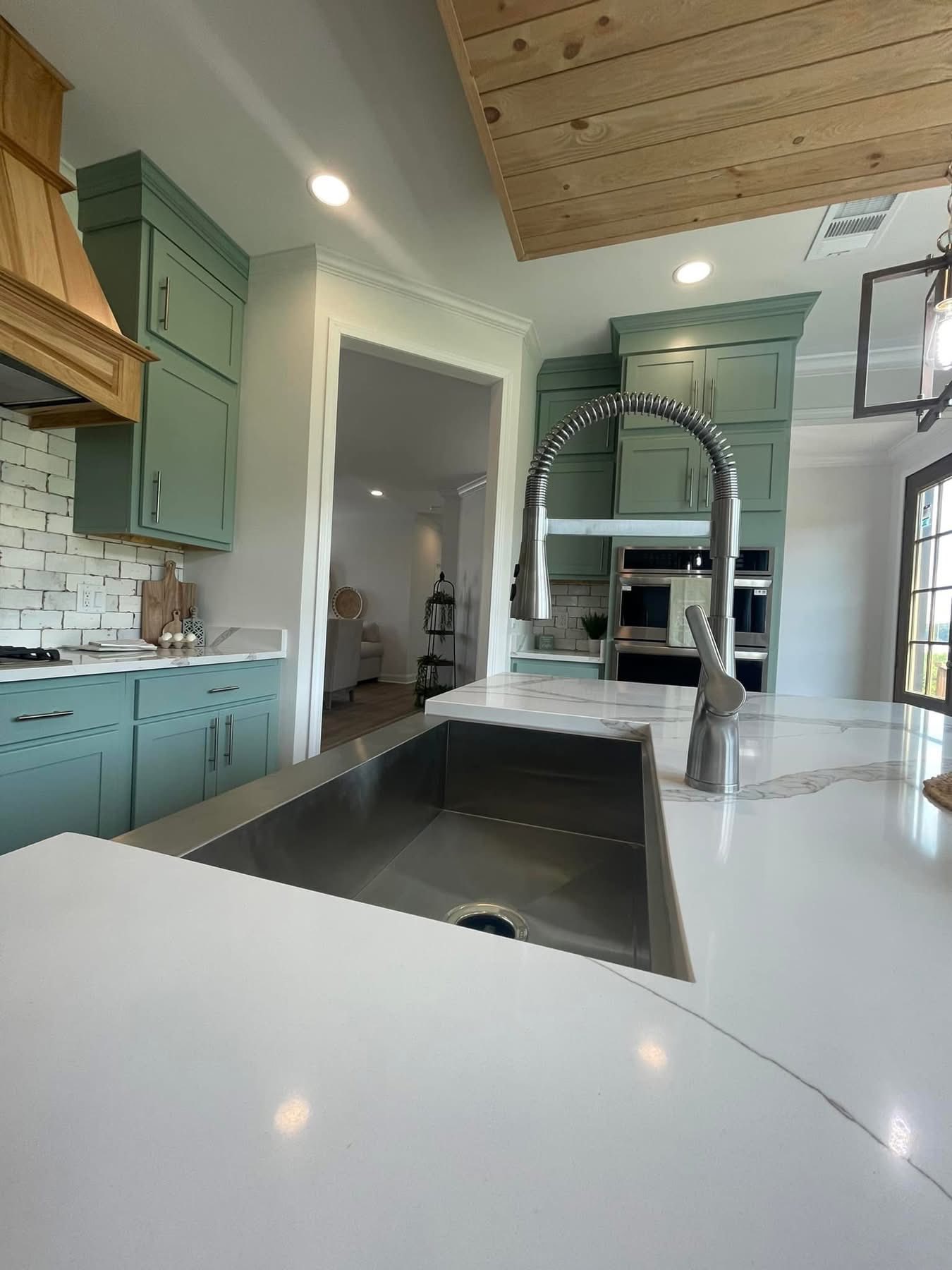 Modern kitchen with green cabinets, a stainless steel sink, and a gooseneck faucet on a white marble countertop. Light wood accents add warmth.