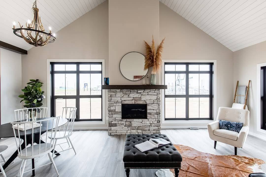 Spacious living room with high ceiling and two large windows. Stone fireplace with round mirror, modern furniture, chandelier, and warm tones. Cozy ambiance.