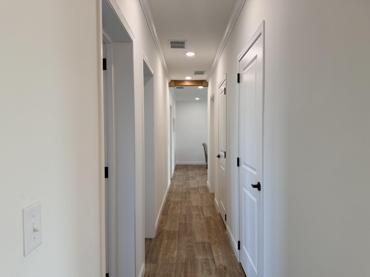 A bright, narrow hallway with light wooden floors, white walls, and several white doors on both sides. Ceiling lights illuminate the space warmly.
