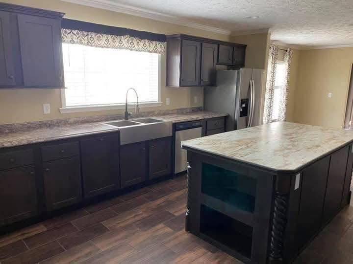 Spacious kitchen with dark cabinets, a large island, farmhouse sink, and stainless steel fridge. Sunlight streams through a window, creating a cozy atmosphere.