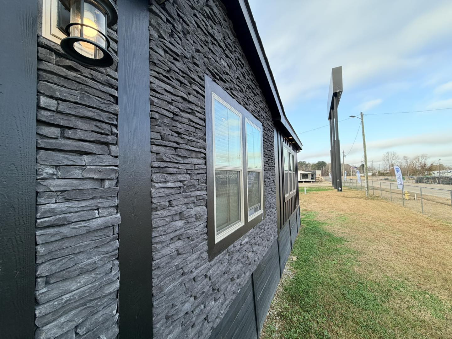 Close-up of a dark stone-textured building wall with two windows reflecting a cloudy sky. A lit lantern is visible; a grassy area leads to a distant road.