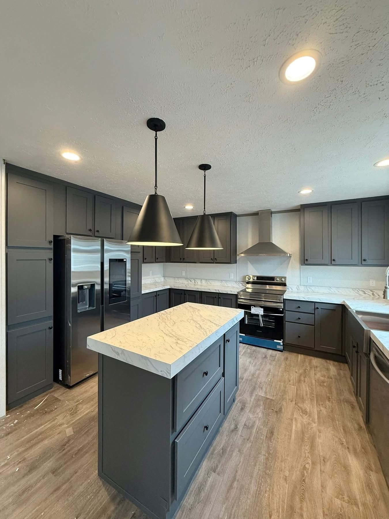 Modern kitchen with grey cabinets and a marble countertop island. Stainless steel appliances, including a fridge and oven. Pendant lights hang overhead.