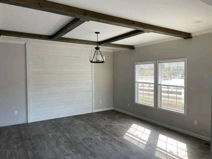 Spacious room with gray hardwood floors, large windows, and a white shiplap accent wall. Exposed wooden beams and a black metal chandelier add rustic charm.