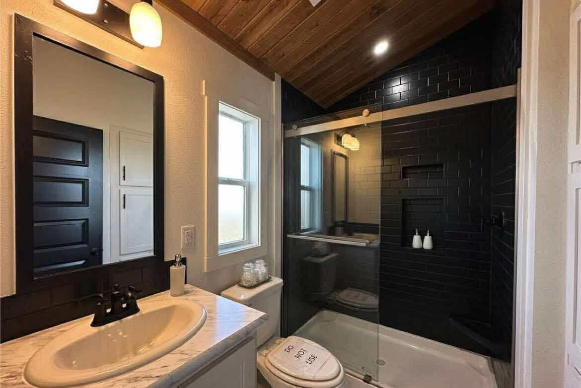 Cozy bathroom with wooden ceiling, black tiled shower, a white sink on a marble counter, mirror, and window. Warm light creates a serene atmosphere.