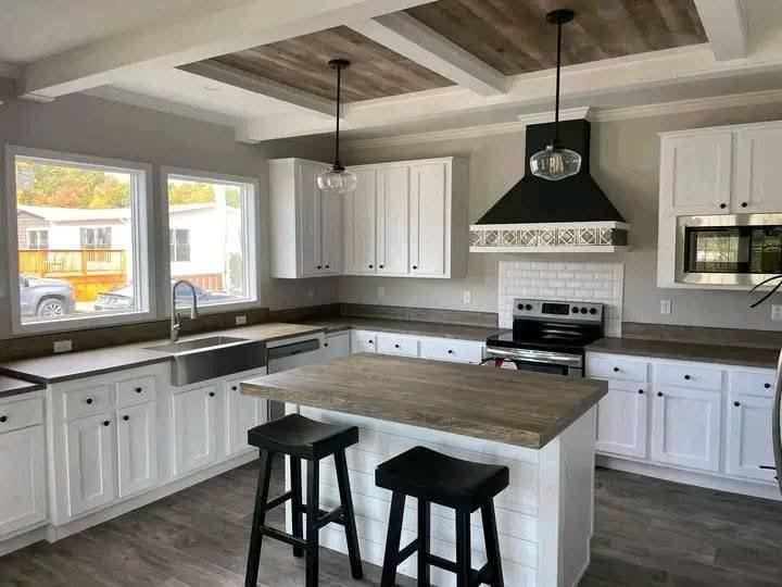 Modern kitchen with white cabinets, wooden island, two black stools, stainless steel appliances, and a decorative hood. Bright and spacious.