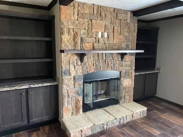 Stone fireplace with a wooden mantel, flanked by dark wooden built-in bookshelves. The room has a cozy, rustic feel with warm earth tones.