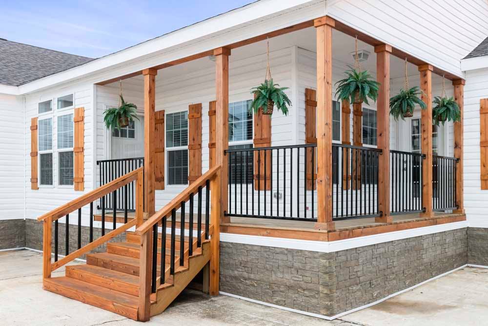 A cozy white house porch with wooden columns, featuring hanging fern plants and black railings. Stone accents at the base enhance charm and elegance.
