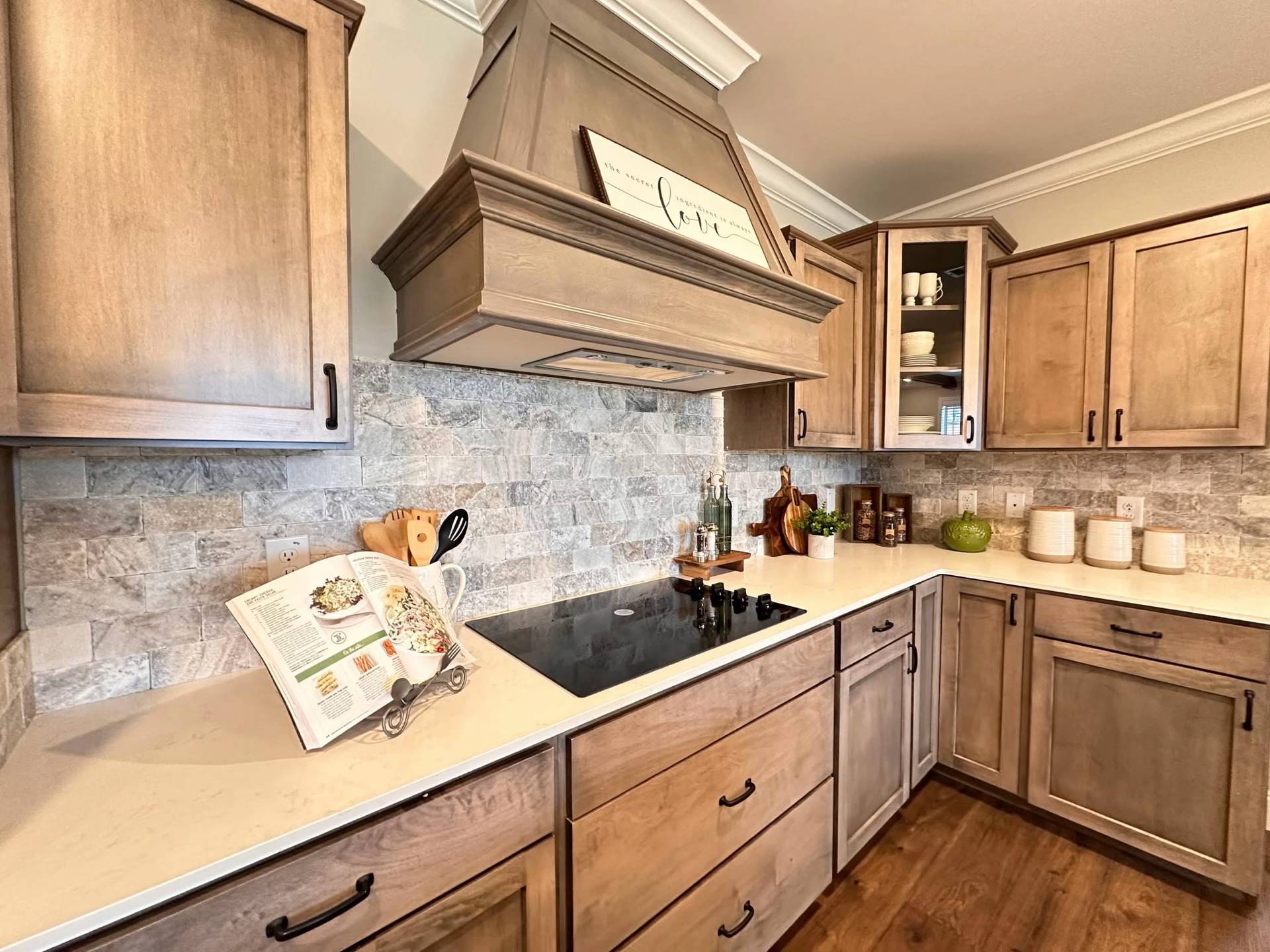 A rustic kitchen with wooden cabinets, stone backsplash, and a modern induction cooktop. An open cookbook and utensils rest on the counter, evoking a cozy and inviting atmosphere.