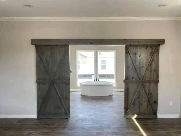 Rustic barn doors slide open to reveal a modern bathroom with a white freestanding tub by large windows, creating a blend of farmhouse and contemporary styles.