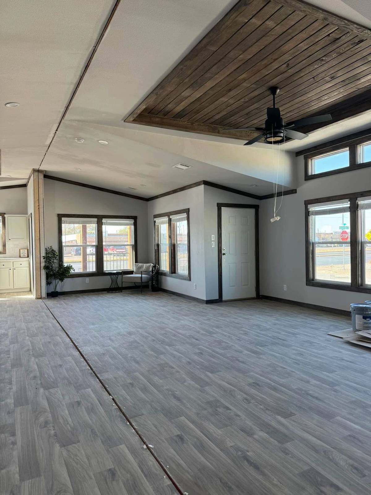 Spacious room with gray wood flooring, a wooden ceiling accent, ceiling fan, large windows, white walls, and a small side table with plants. Bright and modern.