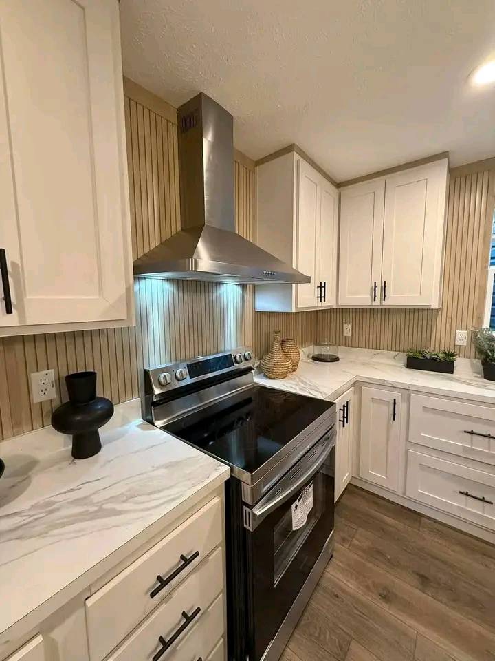 Modern kitchen with white cabinets, black handles, and wood panel backsplash. Features a stainless steel stove and hood, white marble countertops, and decor items.