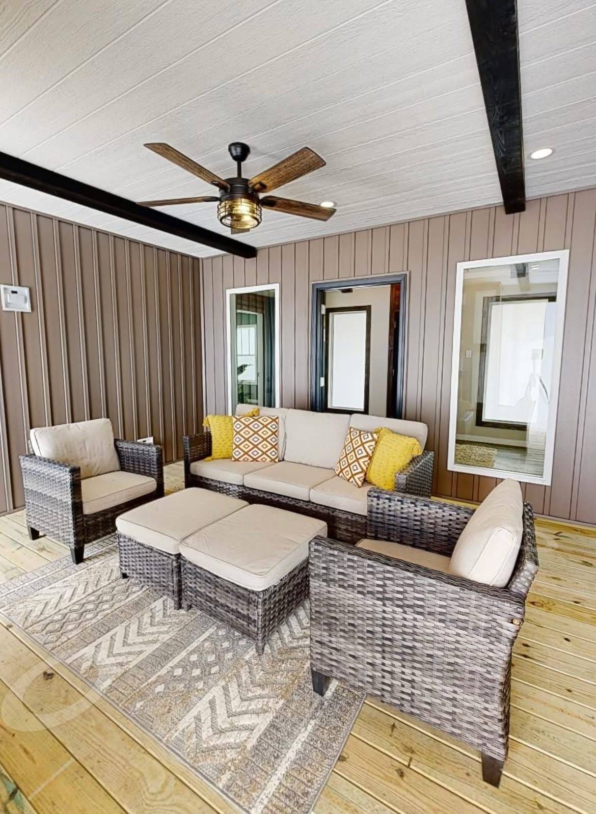 Covered patio with wicker furniture, beige cushions, and yellow pillows. A ceiling fan hangs above, while earth-toned walls add warmth.