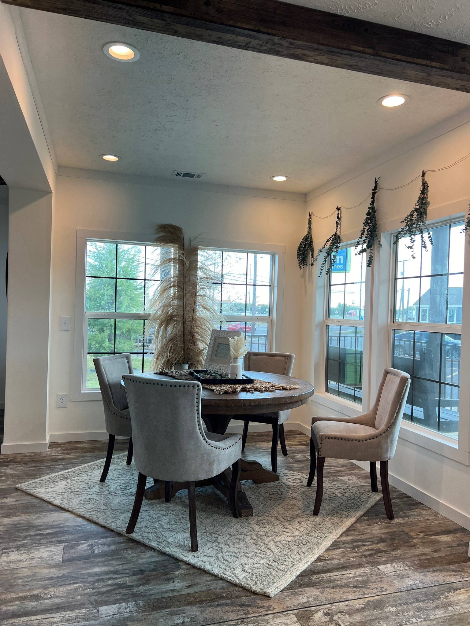 Sunlit dining area with a round table, four upholstered chairs, and pampas grass centerpiece. Large windows with hanging greenery create a cozy, elegant atmosphere.