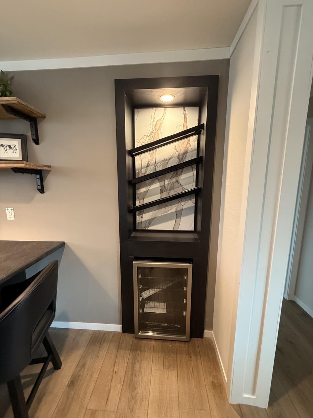 Modern interior with a wine rack in a sleek black frame, set against a marbled backdrop. Below is a small glass-door fridge. Wooden shelves and a dark chair are visible.