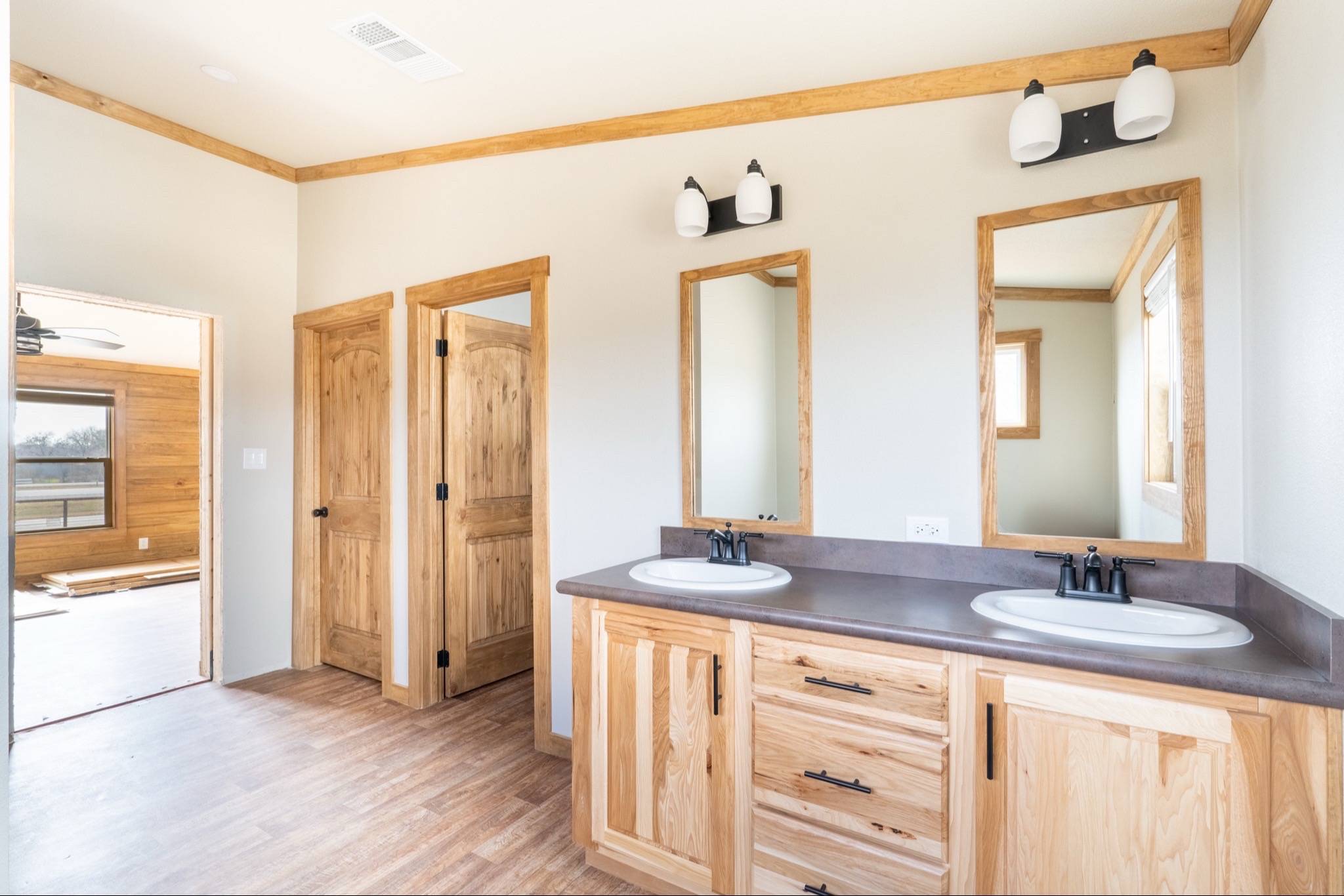 Bright bathroom with two wooden-framed mirrors above dual sinks. Light wood cabinets and doors, alongside tan walls, create a warm, inviting atmosphere.