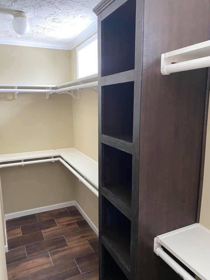 Compact walk-in closet with beige walls, dark wood shelves, white hanging rods, and brown tiled flooring. Brightly lit by a ceiling light.