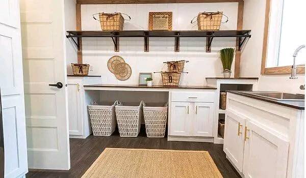 A bright, organized laundry room with white cabinets, a wooden countertop, wicker baskets on a shelf, laundry hampers below, and a beige rug on dark wood floors.