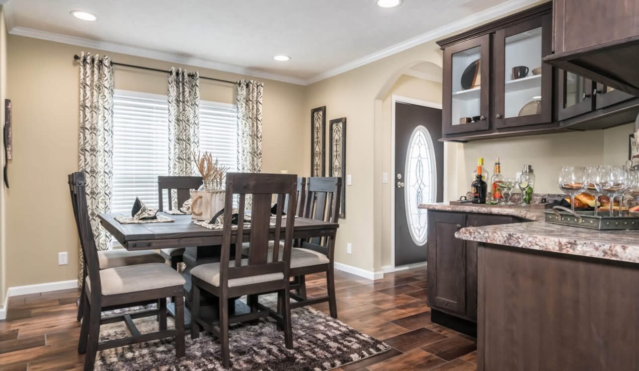 A modern dining room with a wooden table and six chairs on a patterned rug. Curtains with floral designs adorn the windows, and a sideboard holds glassware and bottles. The room has a cozy, inviting ambiance with warm lighting.