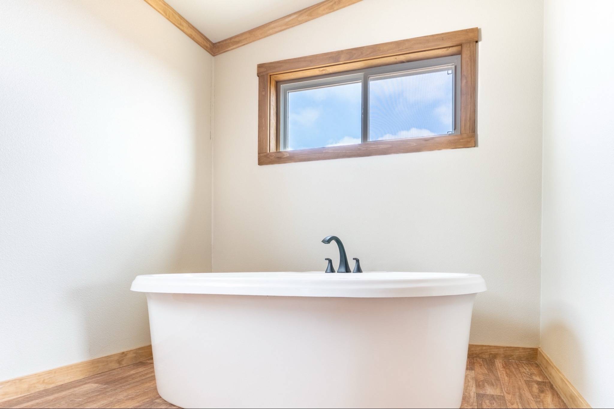 Minimalist bathroom with a white freestanding bathtub, wood floor, and a window with blue sky view. Clean and serene ambiance.