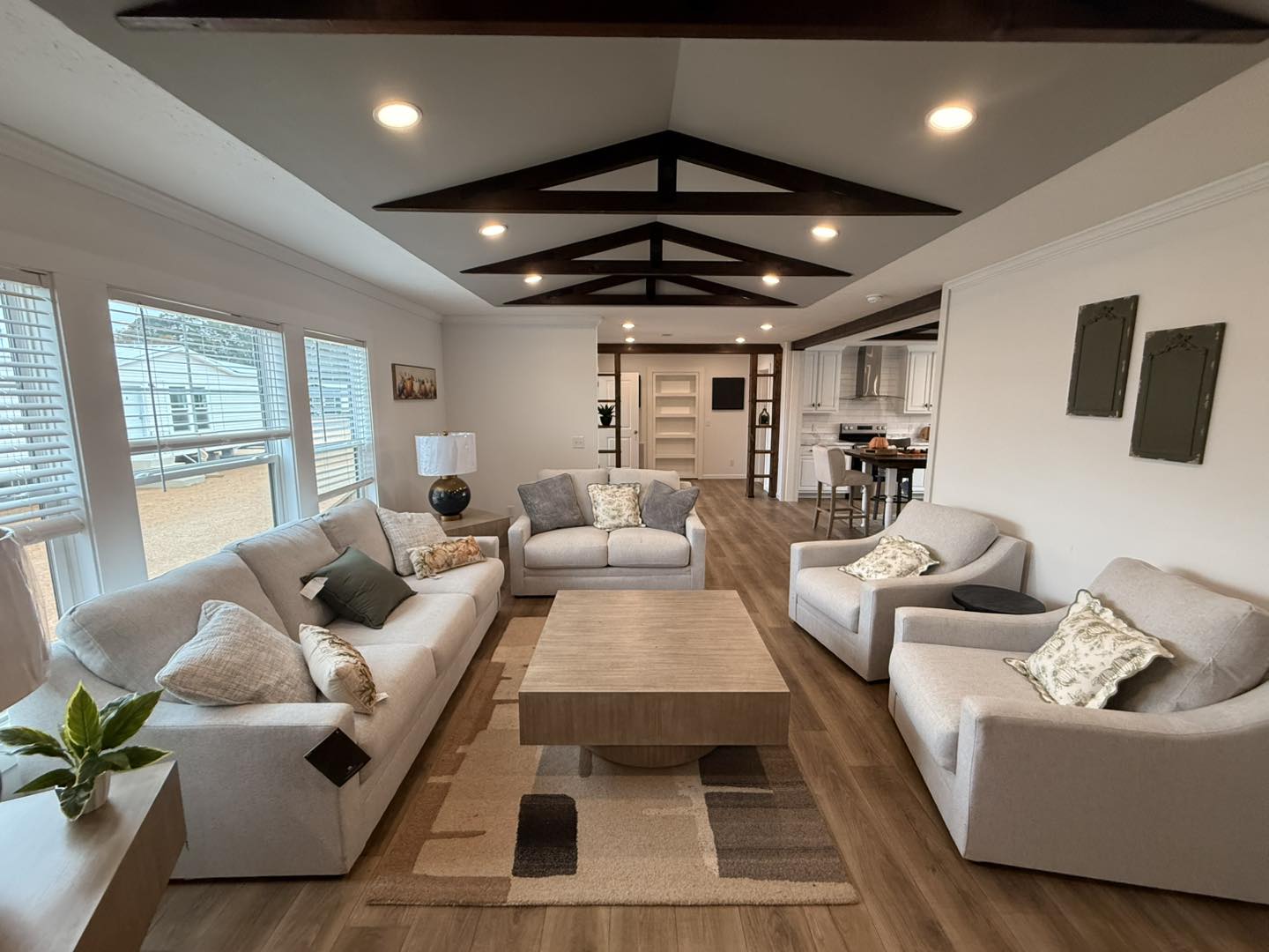 Modern living room with cream sofas around a wooden coffee table on a patterned rug. Large windows, wooden ceiling beams, and open kitchen in the background. Cozy and inviting atmosphere.