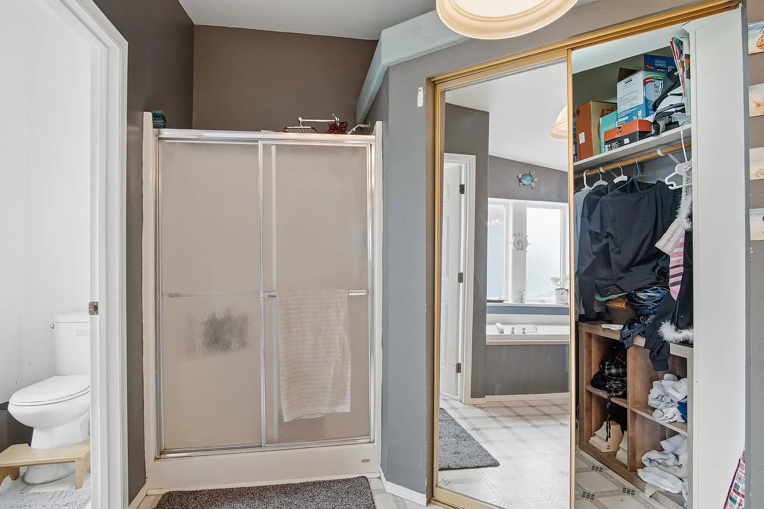 Bathroom with shower, mirrored closet, and a glimpse of a tub in the background. The closet is open, showing clothes and storage boxes on shelves.