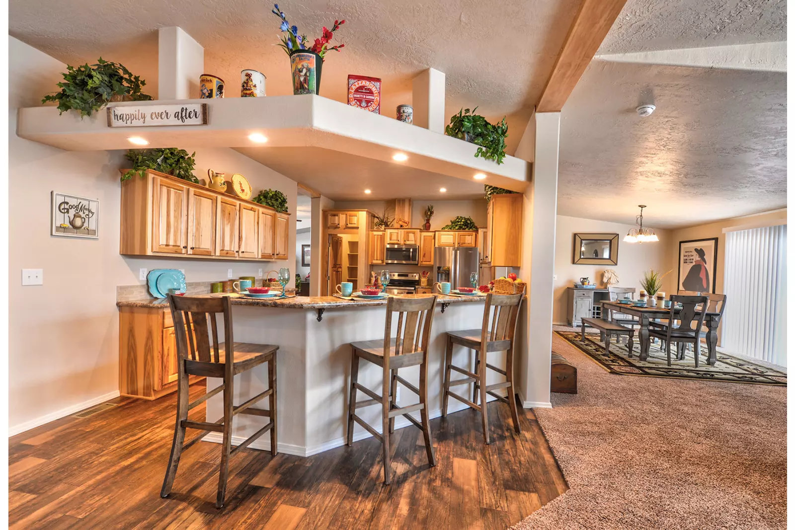 Warm and inviting open kitchen-dining area. Wooden cabinets, bar with three chairs, decor on shelves, plants, and dining table under soft lighting. Cozy ambiance.