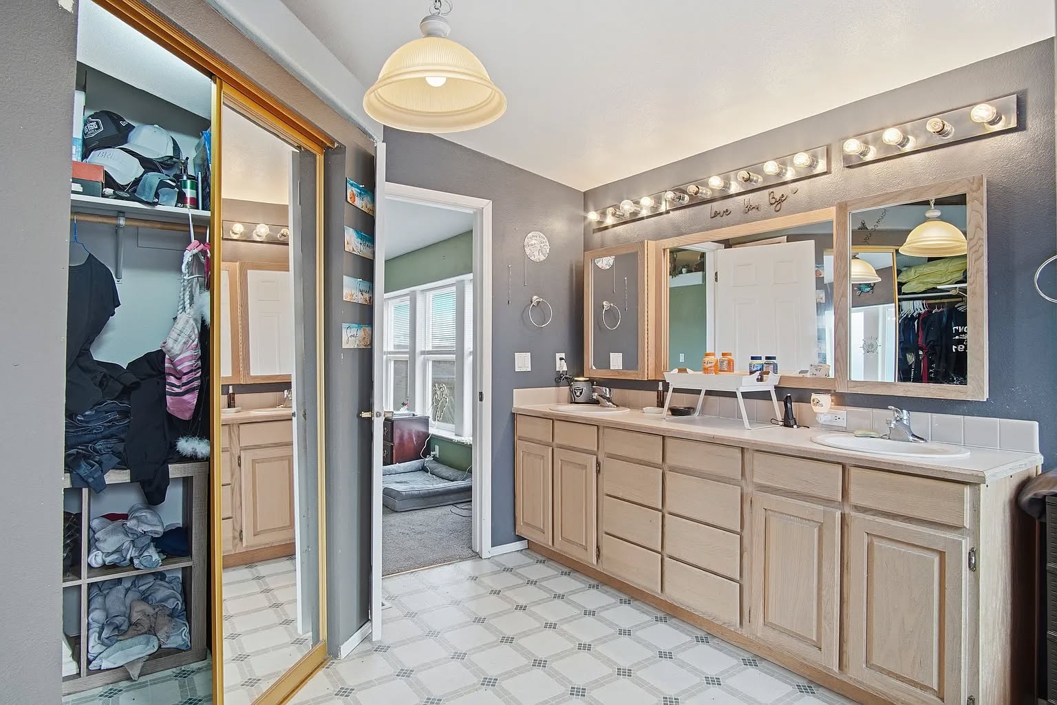 Spacious bathroom with wood cabinets and dual sinks under bright vanity lights. Reflective surfaces include a mirror and sliding closet door, conveying tidiness.