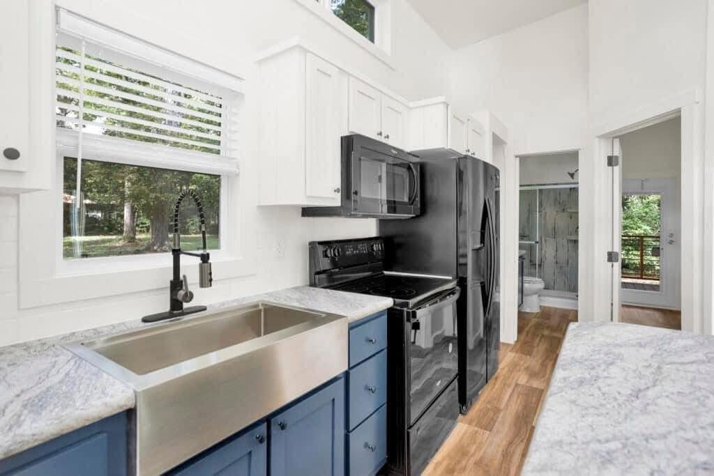 Modern kitchen with blue cabinets, marble countertops, and stainless steel sink. Black appliances include a microwave and fridge. Wood floor and bright window.