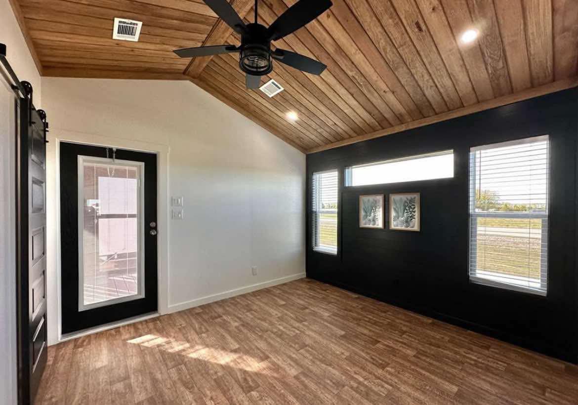 Modern room with a wooden ceiling and floor, black accent wall with art, and large windows. Features a black ceiling fan and a glass door. Bright and airy ambiance.