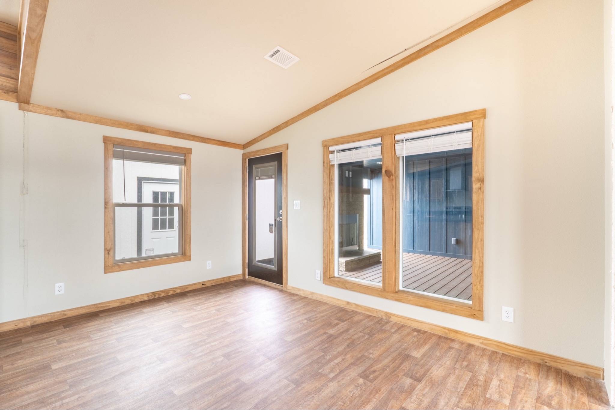 A bright, empty room with light wood flooring and beige walls. It features large windows and a glass door framed with wood, evoking a warm, inviting ambiance.