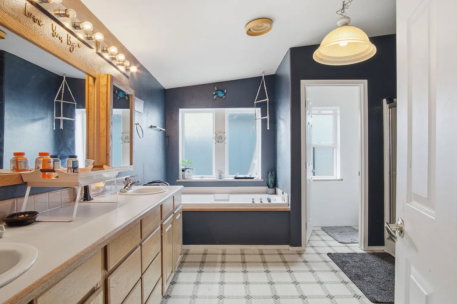 A spacious bathroom with a double sink vanity, large mirror, and warm lighting on the left. Center features a tub by a window, creating a serene atmosphere.