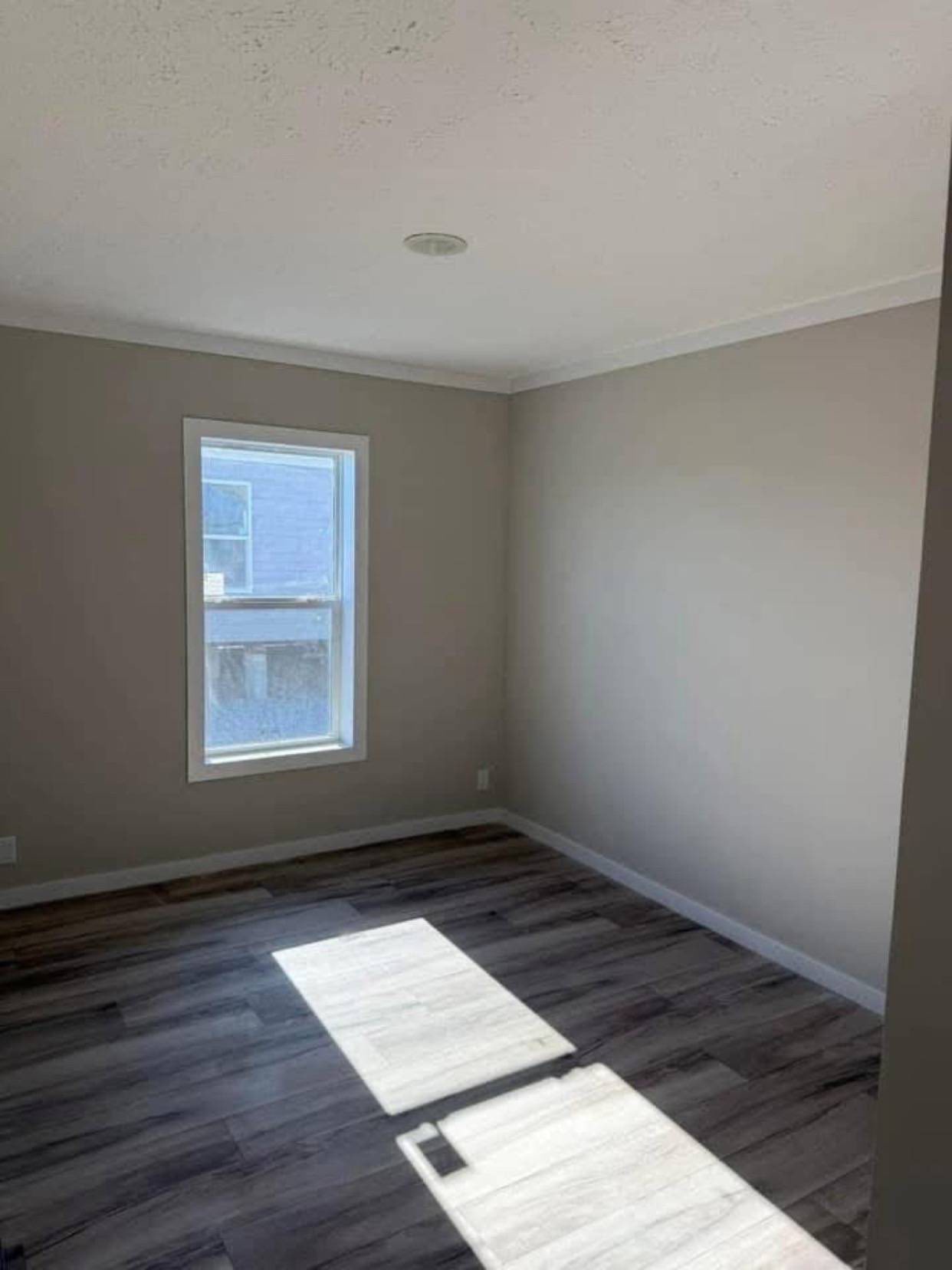 Empty room with light gray walls and a window on the left, letting in sunlight that casts a rectangle on the dark wood floor. Minimalist and bright atmosphere.